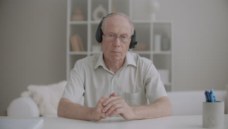 Un-Hombre-Anciano-Está-Escuchando-Una-Conferencia-En-Línea-A-Través-De-Auriculares-Asintiendo-Con-La-Cabeza-Acuerdo-Retrato-Medio-En-Casa
