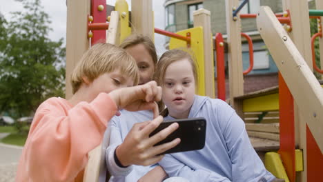 close-up view of a little girl with down syndrome and her friends making a selfie with smartphone in the park on a windy day