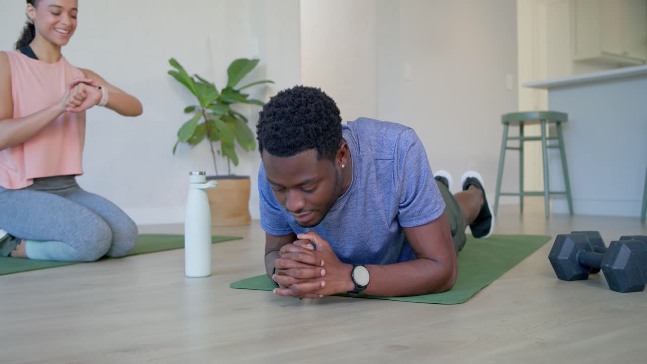 Fitnesspartners doen een onderarmplank op een groene mat thuis, terwijl de vrouw op haar horloge kijkt en de set timed.