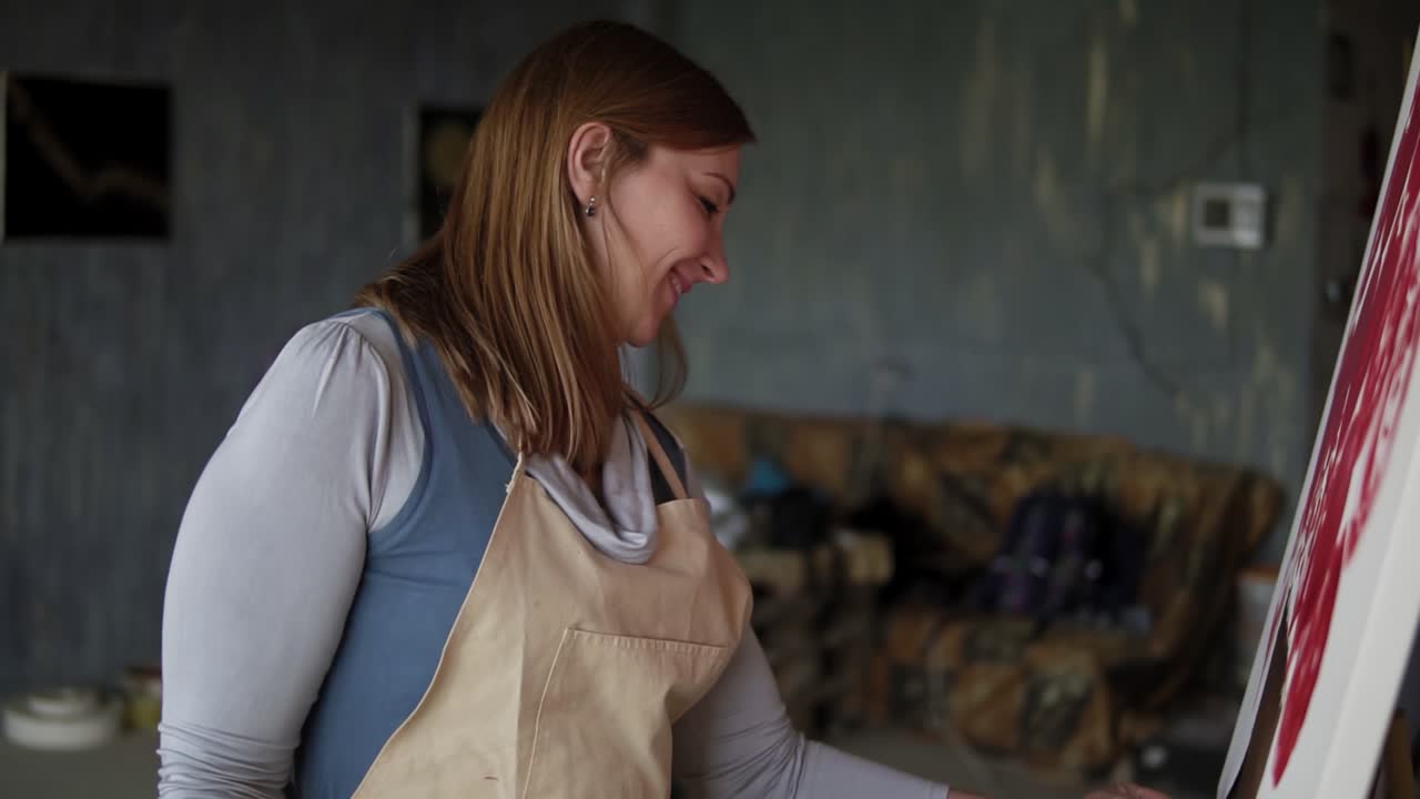 retrato de una mujer caucásica pintando en un estudio de artes usando pinturas de mancha y aceite. artista de cabello largo con delantal beige, sonriendo mientras procesa