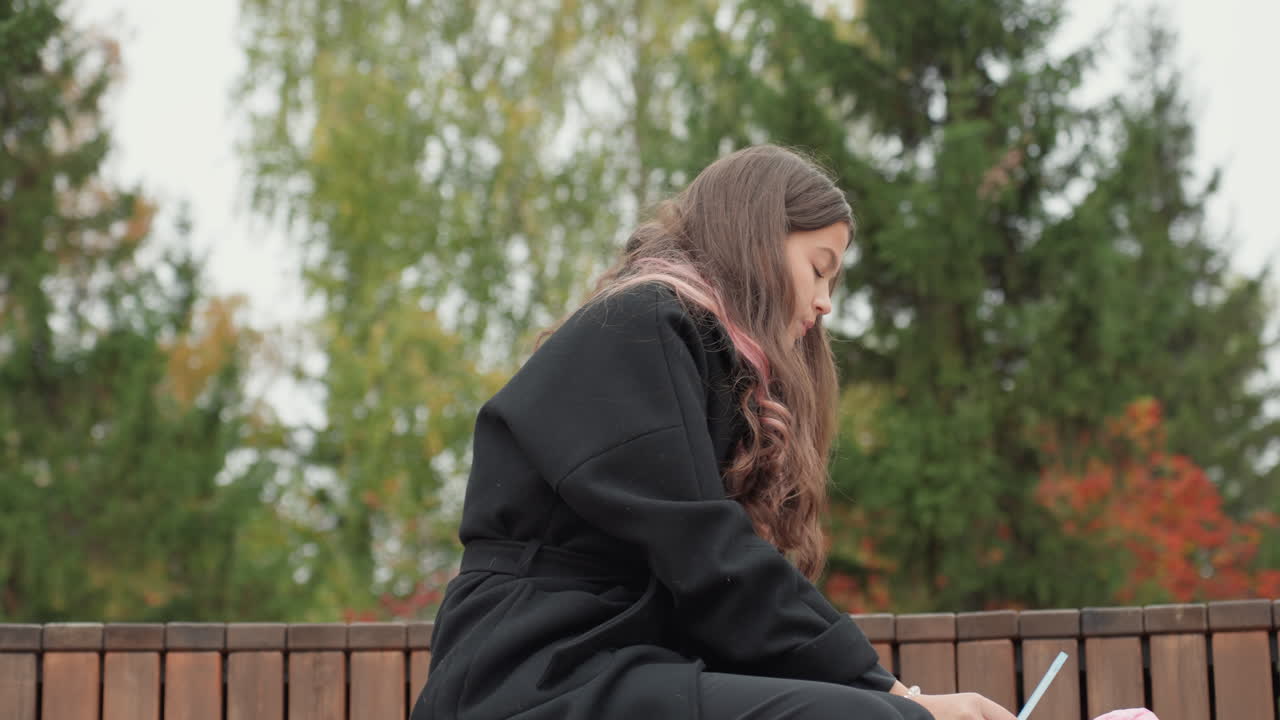 caucasian student studying on bench, autumn campus backdrop with pine trees, overcast sky, dark coat and pinktipped hair, writing in notebook with pen, focused pensive expression, solo outdoor study