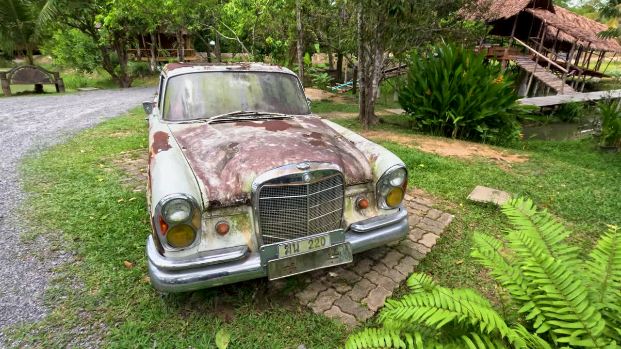 Weathered antique car sits near lush greenery and rustic wooden house, daylight, slight camera movement