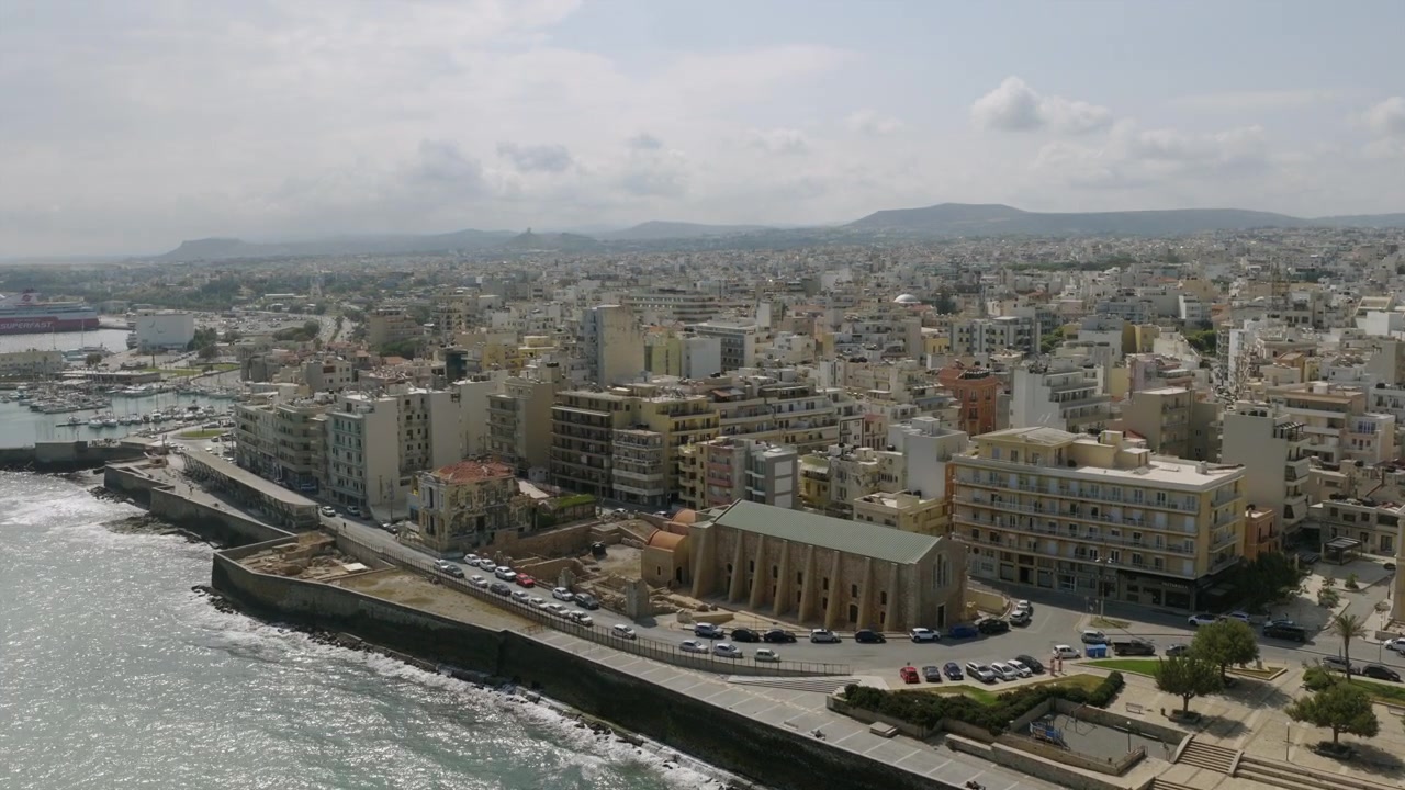 Veduta aerea del lungomare di Heraklion a Creta, con onde che si infrangono lungo la diga foranea e densi edifici cittadini che si ergono verso distanti montagne velate