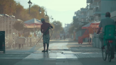 Man-cleaning-asphalt-road-next-to-street-cafe-with-hose