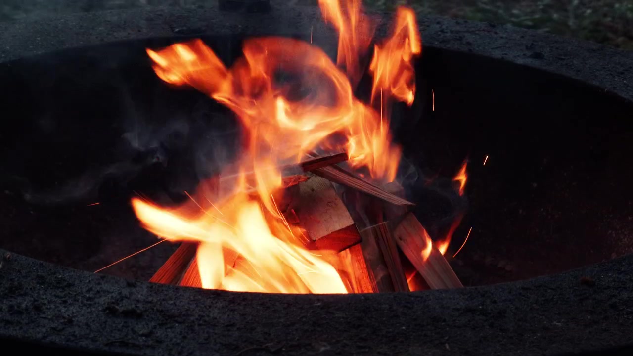 Legna che brucia all'interno di un braciere da esterno con fiamme arancioni brillanti, fumo e piccole scintille. Primo piano di un caldo falò che arde la sera.