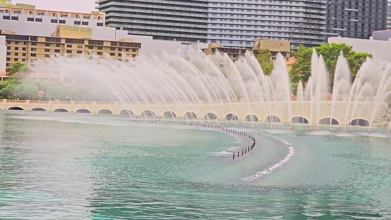 Espectáculo de fuentes del Strip de Las Vegas en el Bellagio, con altos chorros de agua que se elevan del lago, torres de hoteles, árboles, arcos de piedra y brillantes reflejos diurnos en el agua turquesa