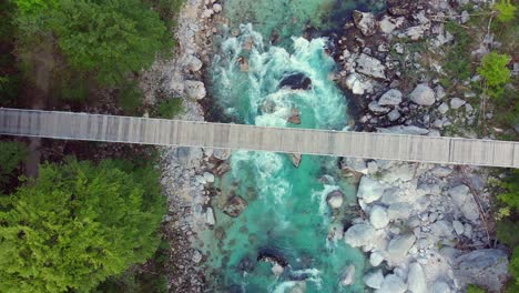 drone spirals down toward footbridge, wild mountain river flows below