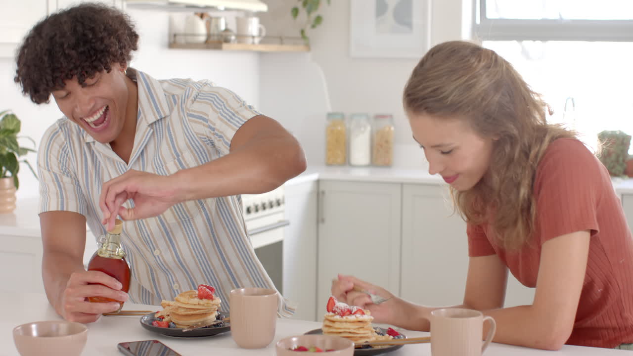 Young diverse couple enjoying breakfast, pouring syrup on pancakes at home kitchen table