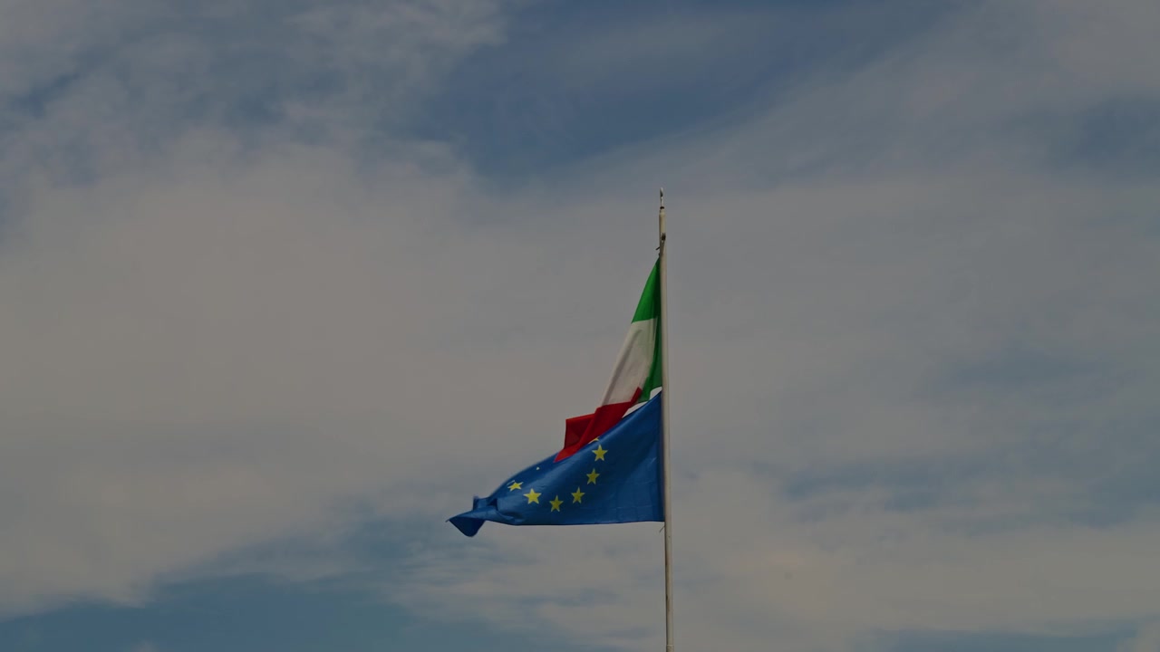 Bandiere dell'Italia e dell'Unione Europea sventolano con grazia nel vento contro un cielo azzurro limpido con nuvole soffici. Simbolo di unità europea, identità e orgoglio nazionale