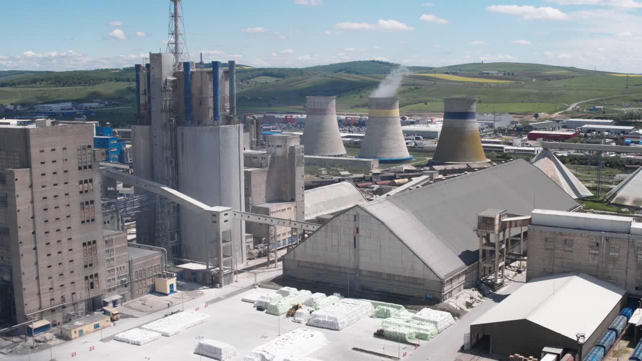 A wide aerial drone shot orbits a massive industrial complex. The view shows concrete silos, smokestacks, and large cooling towers emitting steam, with a rural landscape behind