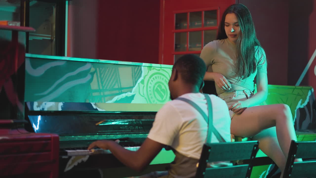 un hombre negro toca el piano para su novia europea en un bar. una joven seductora escucha música sentada junto a un músico en un club semi-oscuro. una cita romántica de pareja