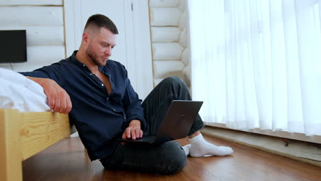 focused bearded man wearing blue shirt sits on the floor at the big bed. man looks at the laptop on his knees.