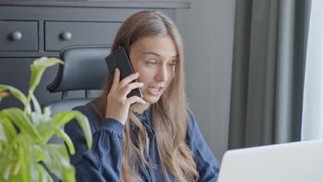 Mujer-Joven-Que-Parece-Sorprendida-Mientras-Habla-Por-Teléfono-Mientras-Trabaja-Con-Una-Computadora-Portátil-En-La-Oficina