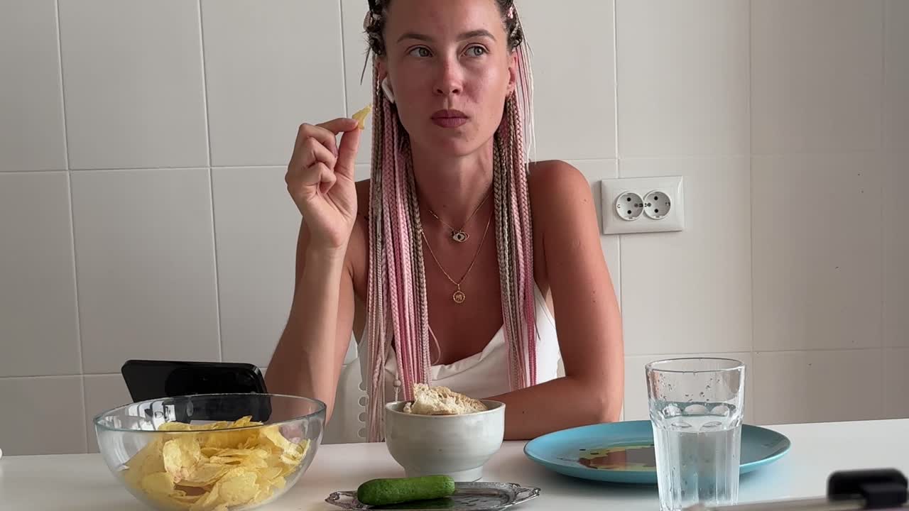 Mujer joven con trenzas rosas comiendo patatas fritas en una mesa de cocina.
