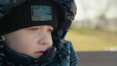 close-up of a child wearing a black jacket and beanie, gazing thoughtfully with a calm expression, with blurred landscape of trees