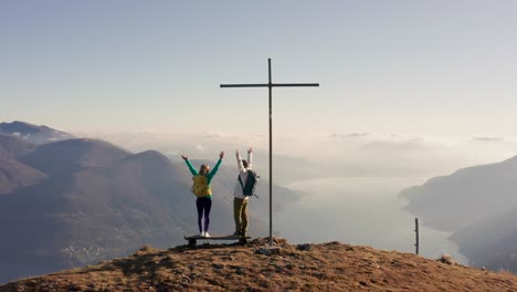 aerial: couple hikes stand on mountain top arms wide open.