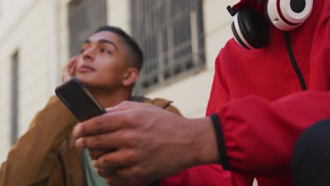 two happy mixed race male friends sitting and using smartphone in the street