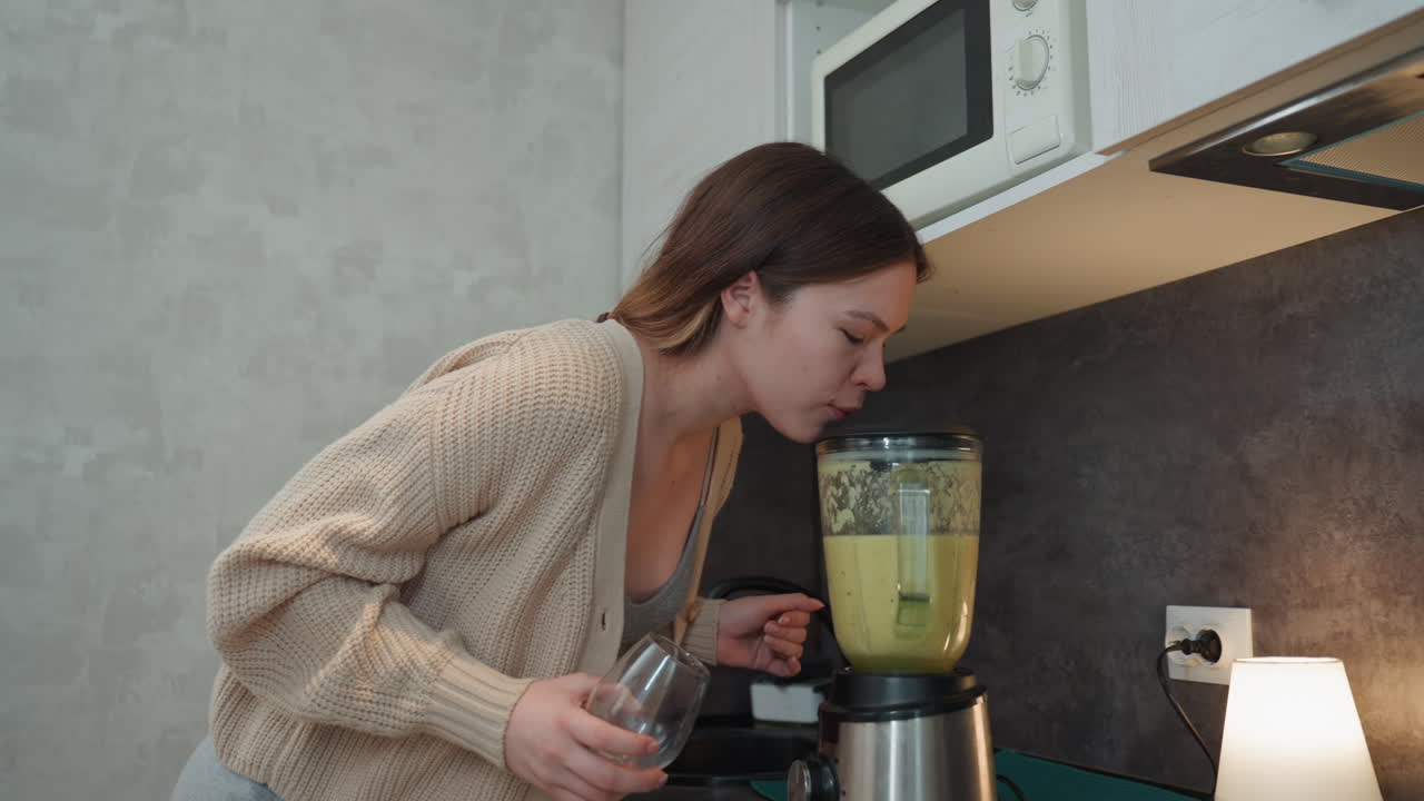 Donna caucasica incinta prepara un frullato verde sul bancone della cucina, tenendo un misurino e un bicchiere, versando gli ingredienti nel frullatore, chinandosi per controllare l'aroma, cardigan accogliente, microonde sopra, caldo