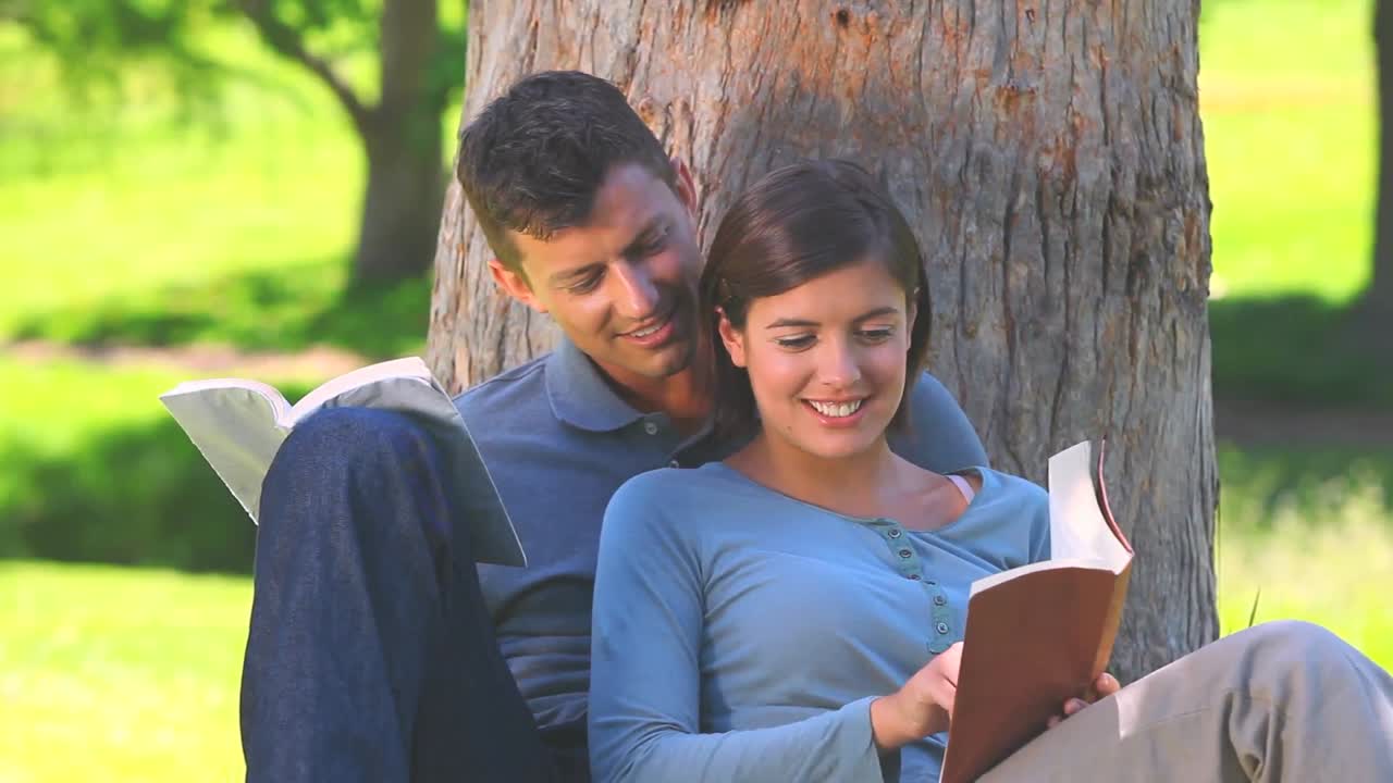 Pareja joven leyendo libros bajo un árbol.