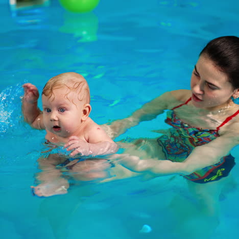 brunette woman holding a tiny baby in the swimming pool. adorable child splashing water cheerfully.