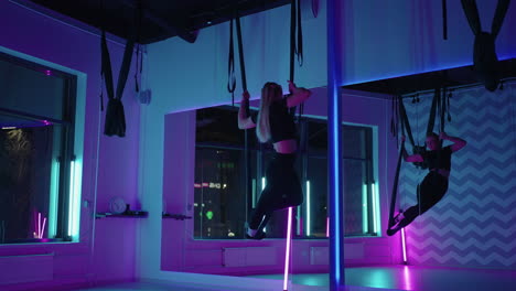 a woman does sports yoga in the air on a hammock in neon light performing graceful exercises and movements from stretching and dancing in slow motion.