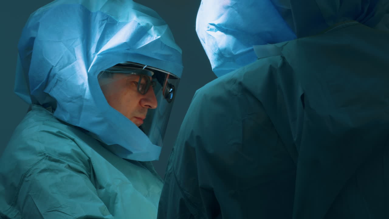 Due chirurghi in cuffie protettive sterili azzurro chiaro che operano sotto una luce intensa. La vista ravvicinata mostra l'intensa concentrazione del team chirurgico durante la procedura.