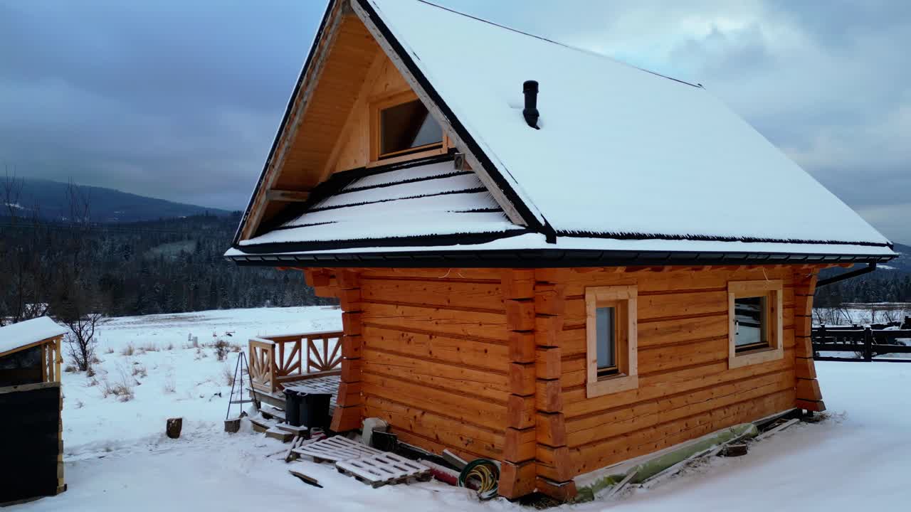 La camera ruota attorno a una baita rustica coperta di neve. Il paesaggio invernale, le montagne boscose e l'atmosfera serena creano una scena alpina cinematografica