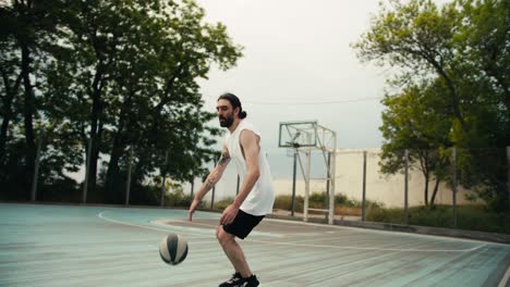 un hombre con una camiseta blanca y pantalones cortos negros dribla hábilmente una pelota de baloncesto en una cancha de baloncesto afuera en verano. deportes de ocio activos al aire libre