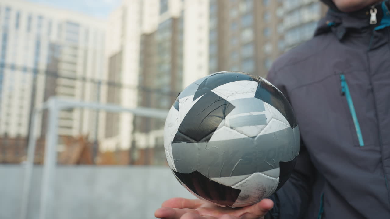hombre habilmente gira la pelota de fútbol en su palma, mostrando equilibrio y enfoque, con edificios urbanos de gran altura y valla de malla en el fondo