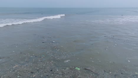 low altitude pan down drone shot of polluted ocean filled with floating trash and debris and muddy sewage river runoff with surfers in the background in bali indonesia