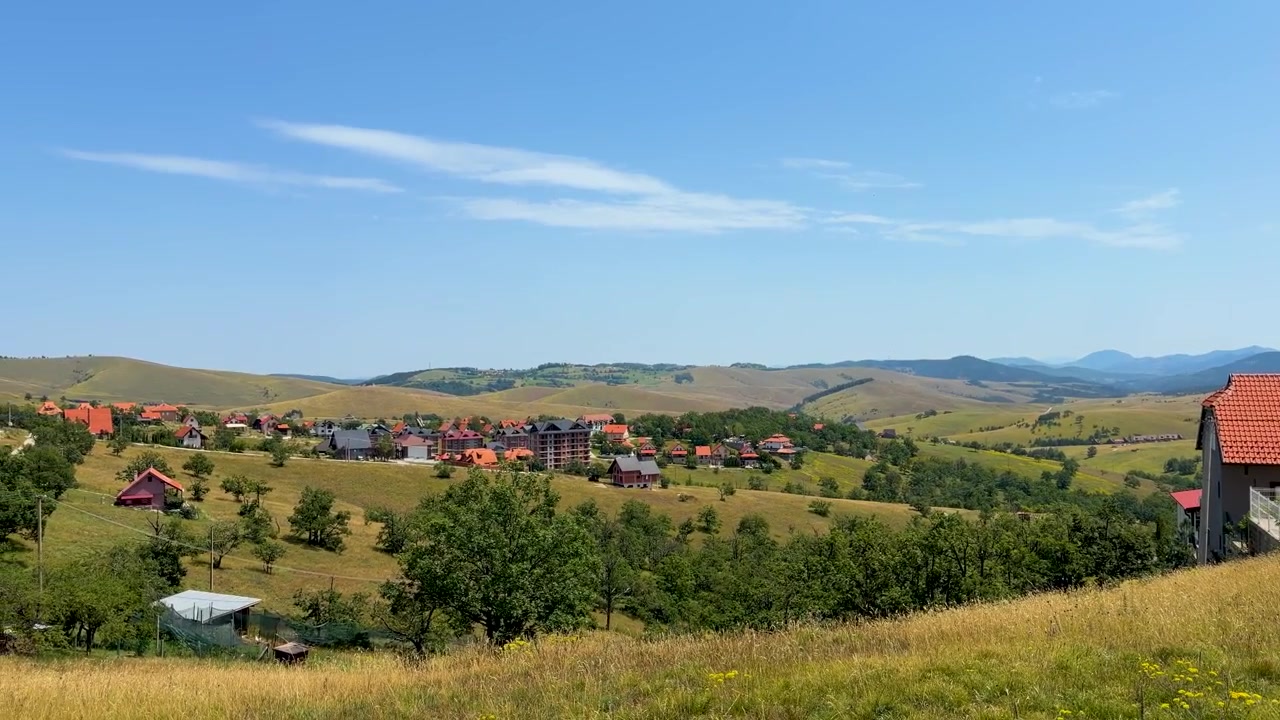 Kamera snímá domy na svahu, travnaté louky, roztroušené stromy a zvlněný horský terén pod jasnou modrou letní oblohou v Zlatiboru, Srbsko.