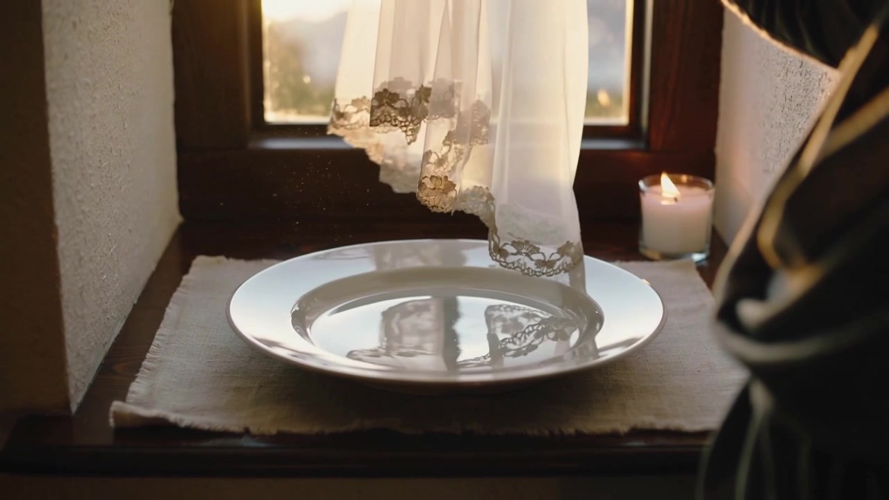 Moving in person wearing dark sleeves lifting lace curtain at sill, showing white plate, lit candle