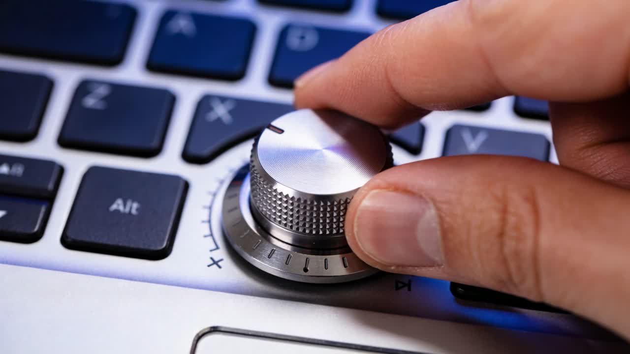 Reaching right hand grasping metallic rotary knob on laptop keyboard, turning dial to adjust volume