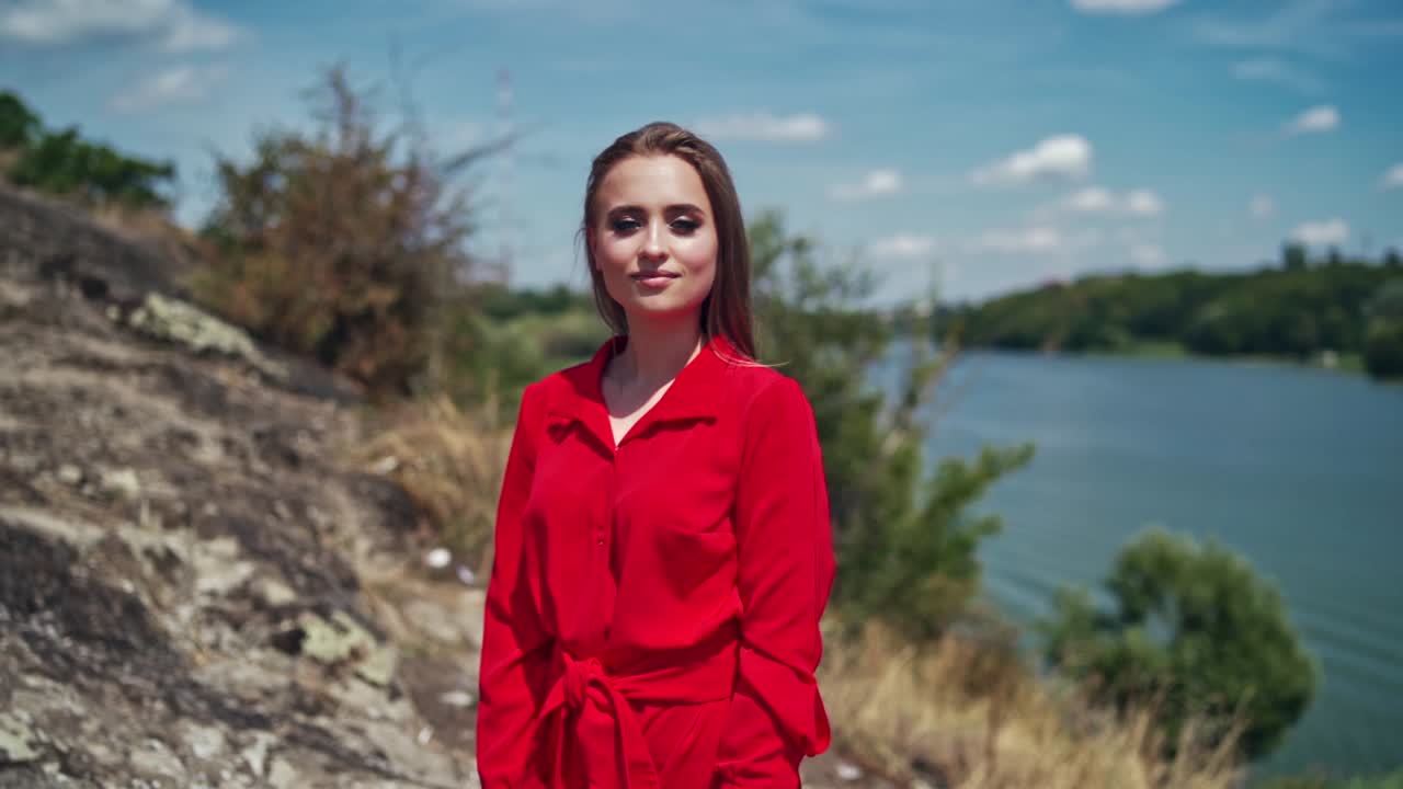 Portrait of a beautiful woman on nature background. Attractive young model in red dress with long hair standing near the river and smiling.