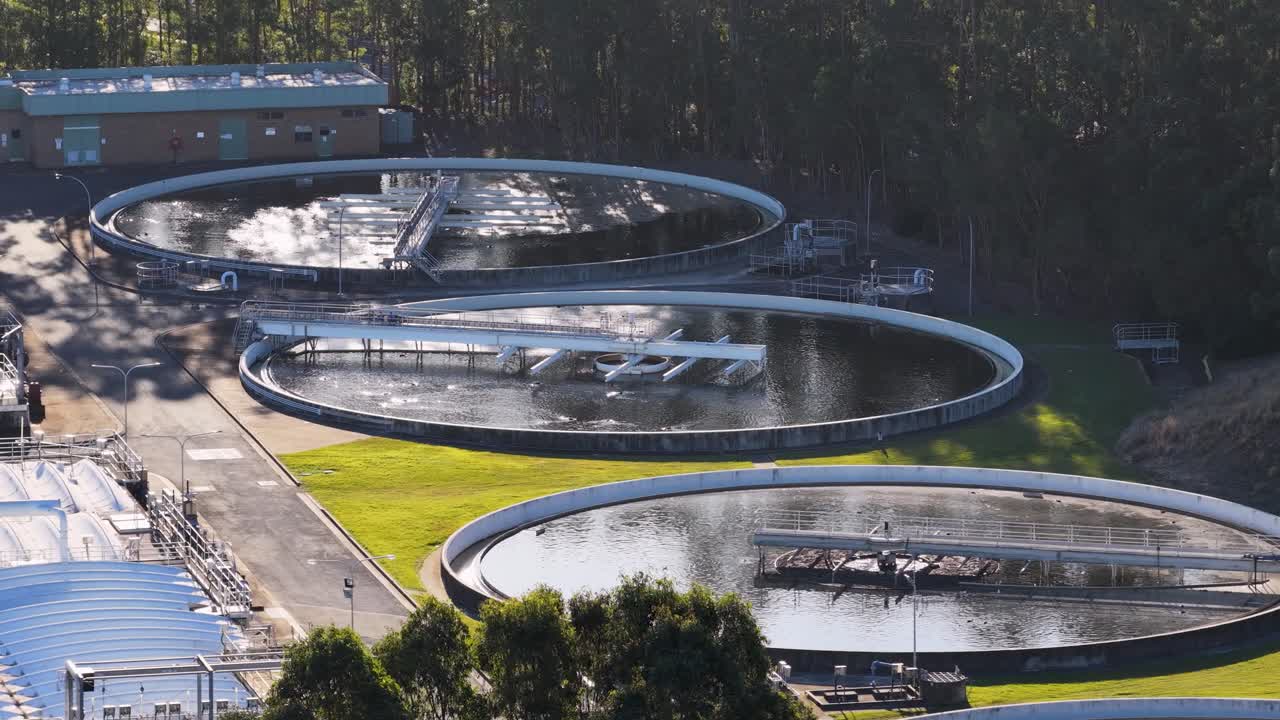 Drone footage pans over circular wastewater treatment tanks surrounded by green lawns and trees, with soft sunset lighting and clear environmental details