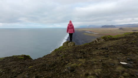 female standing on edge of the cliff at dyrhólaey viewpoint in iceland - aerial pullback