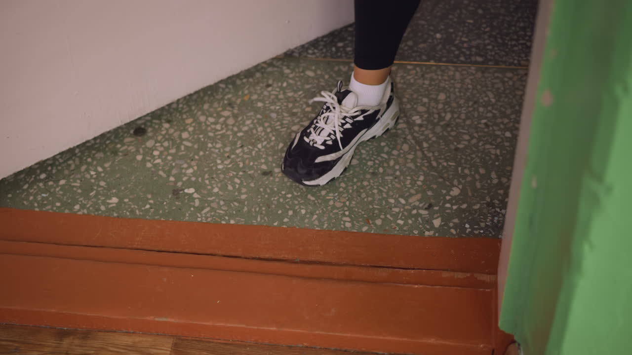 Leg view of girl in black leggings wearing canvas sneakers stepping out from door onto polished wooden ground with green wall and terrazzo floor background showing casual movement