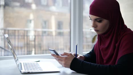 Young-suposedly-muslim-girl-with-purple-hijab-on-her-head-writes-something-down-from-her-smartphone,-sitting-in-a-light-indoor-space-with-glass-windows-on-the-background