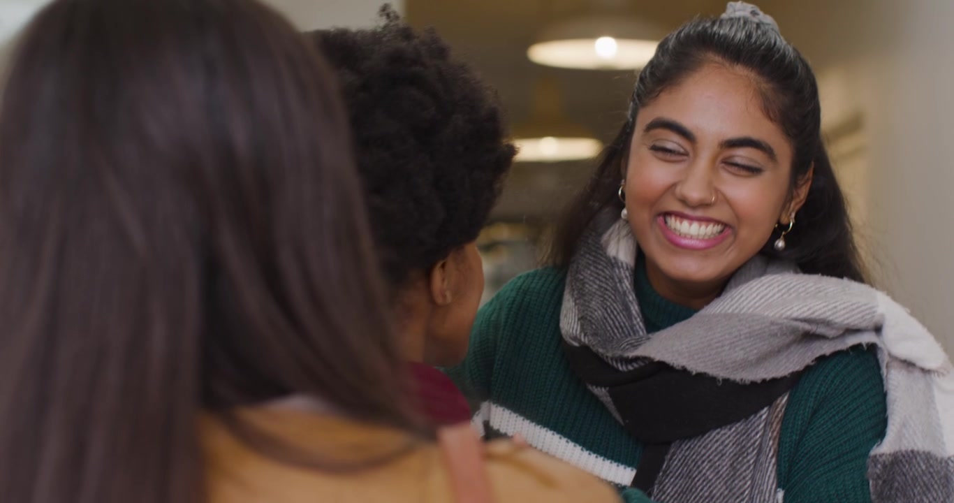 Diverse amiche si salutano nell'atrio, mentre una donna indiana con un maglione entra e le abbraccia.