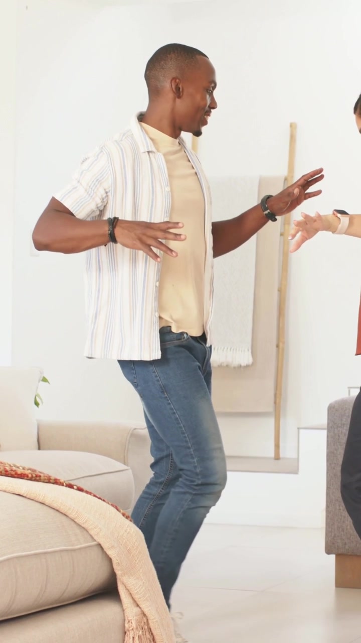 Vertical video: Entering left, man in tee moving across home to woman near sofa gesturing for dance