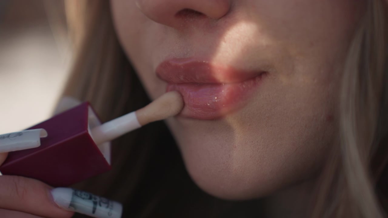 Bella donna che applica il lucidalabbra in morbida luce esterna, primo piano di labbra e applicatore, delicate pennellate, ombre calde sulla guancia, routine di trucco naturale con sorriso sottile e finitura rugiadosa.