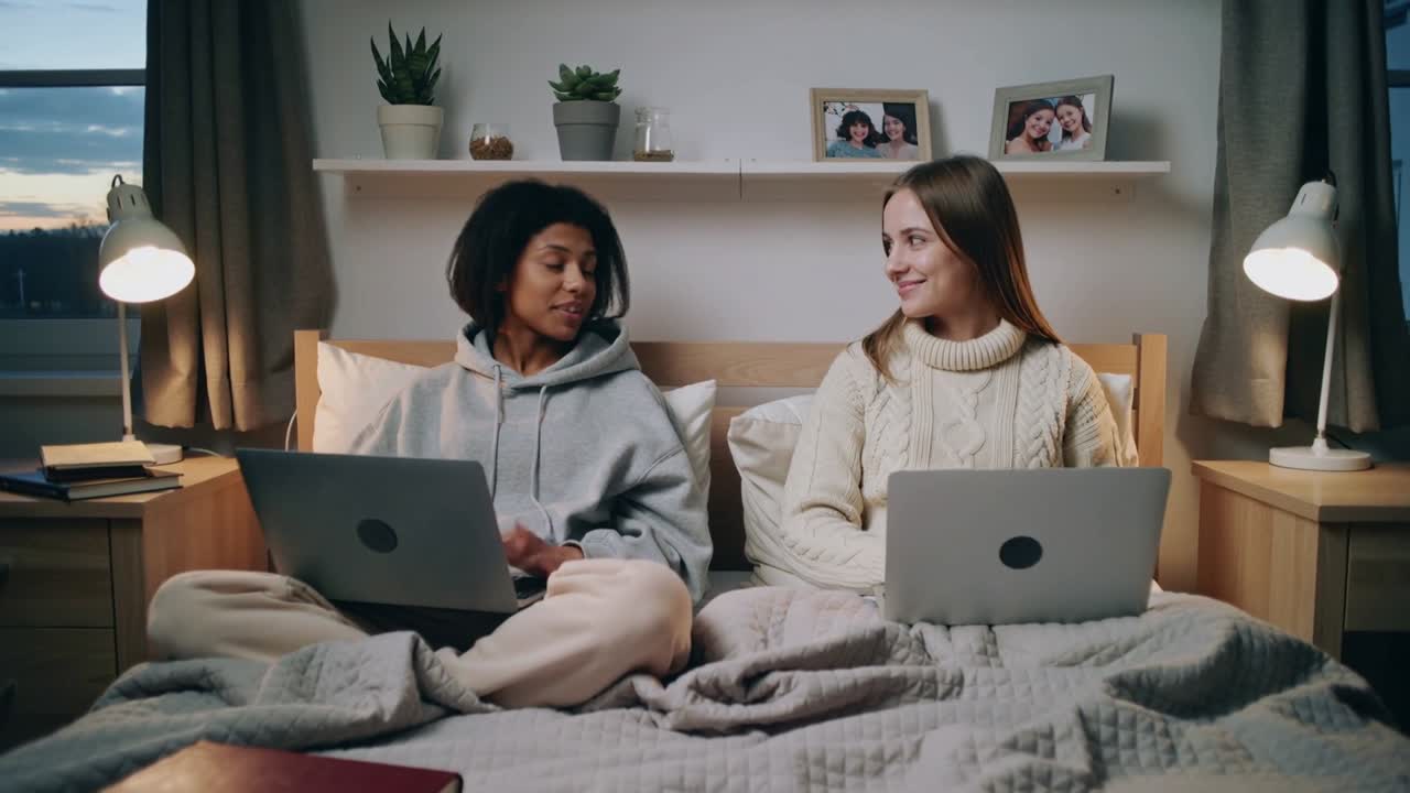 Mirando hacia la izquierda, dos estudiantes con sudadera con capucha y de punto se intercambian sonrisas mientras estudian en la cama con computadoras portátiles.