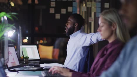 video of tired african american businessman working at desk at night with colleagues in office