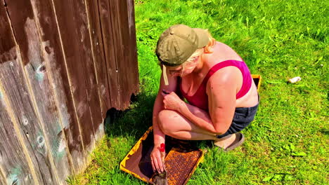 Woman repainting wooden wall with brown paint while kneeling on grass in backyard sunshine