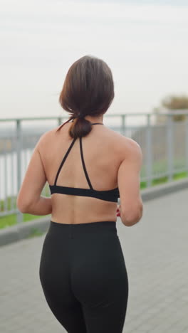 back view of young girl in black neck top and tight leggings jogging in park with blurred background, another woman walking toward her along the park path near iron railing and river