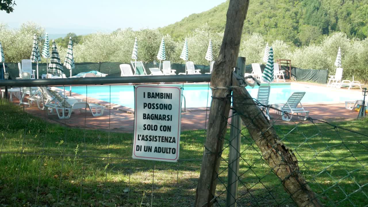 pan a la izquierda de una piscina pública con un cartel de valla que dice "los niños sólo pueden bañarse con la ayuda de un adulto" en italiano