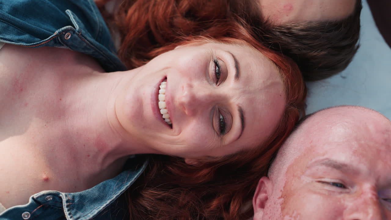 Aerial view of beautiful mother lying upwards between husband and child with eyes closed, smiling peacefully under sunlight, expressing warmth, tenderness, and relaxed family connection