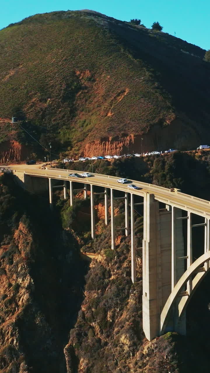 Busy road around the rugged mountain passing into the beautiful arched bridge over the gap in the rocks. Blue skies at backdrop. Top view. Vertical video