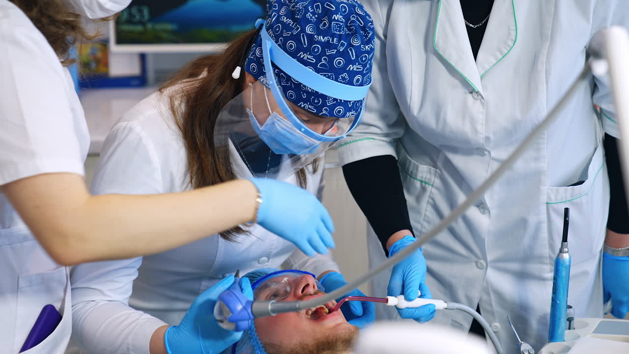 Dentistry student in mask and face shield sits over the patient. Teacher standing beside gives advice and recommendations.