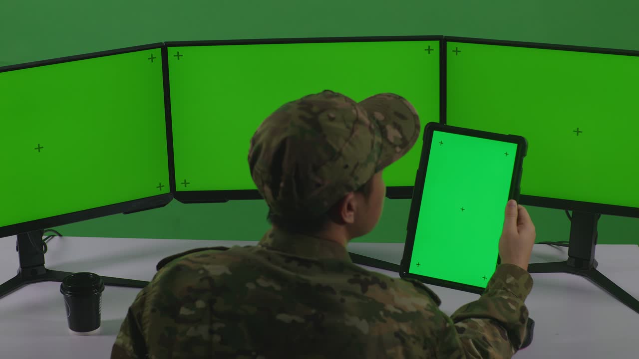 태블릿과 여러 컴퓨터 모니터로 작업하는 아시아 남성 군인의 뒷면으로 녹색 스크린 스튜디오에서 모의 녹색 화면